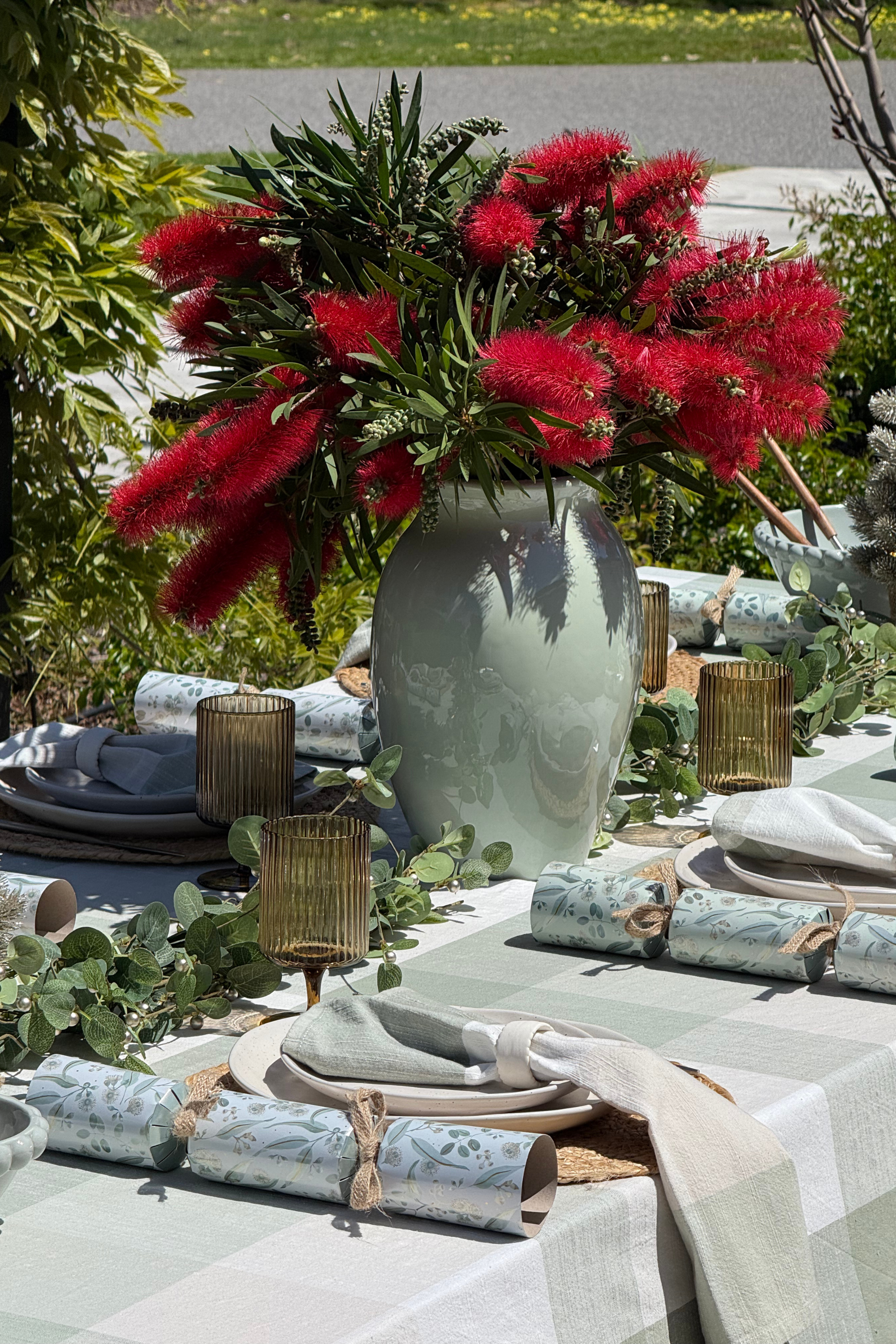 Decorative table setting with red flowers in a vase, greenery, and patterned tablecloth.