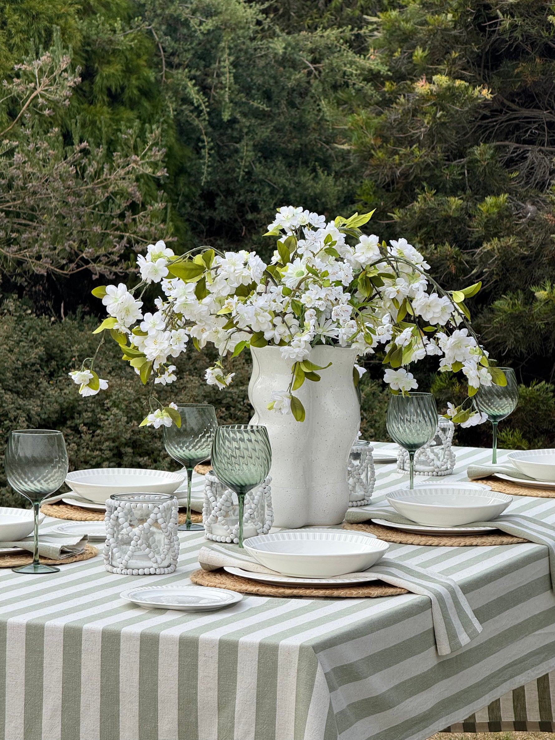 Outdoor table setting with white flowers in a vase, glasses, and plates on a striped tablecloth.