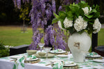 An outdoor table setting styled with a large white ceramic vase filled with white flowers and green leaves, alongside matching green and white patterned napkins, plates, and bowls on a white tablecloth. A lush background of green grass and hanging purple wisteria flowers is visible.