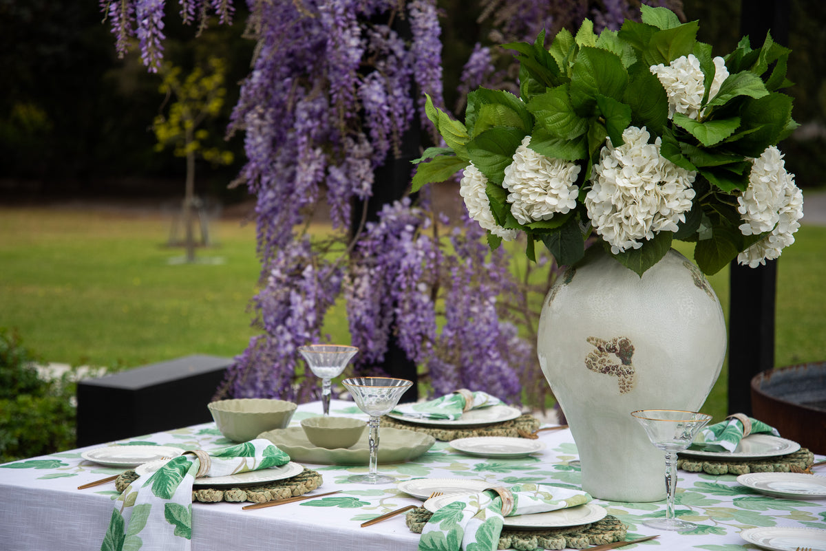 An outdoor table setting styled with a large white ceramic vase filled with white flowers and green leaves, alongside matching green and white patterned napkins, plates, and bowls on a white tablecloth. A lush background of green grass and hanging purple wisteria flowers is visible.