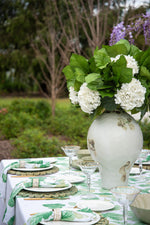 An outdoor garden table setting featuring a large white ceramic vase filled with white hydrangeas and green leaves, styled with a white tablecloth, green napkins, woven placemats, and clear martini glasses.