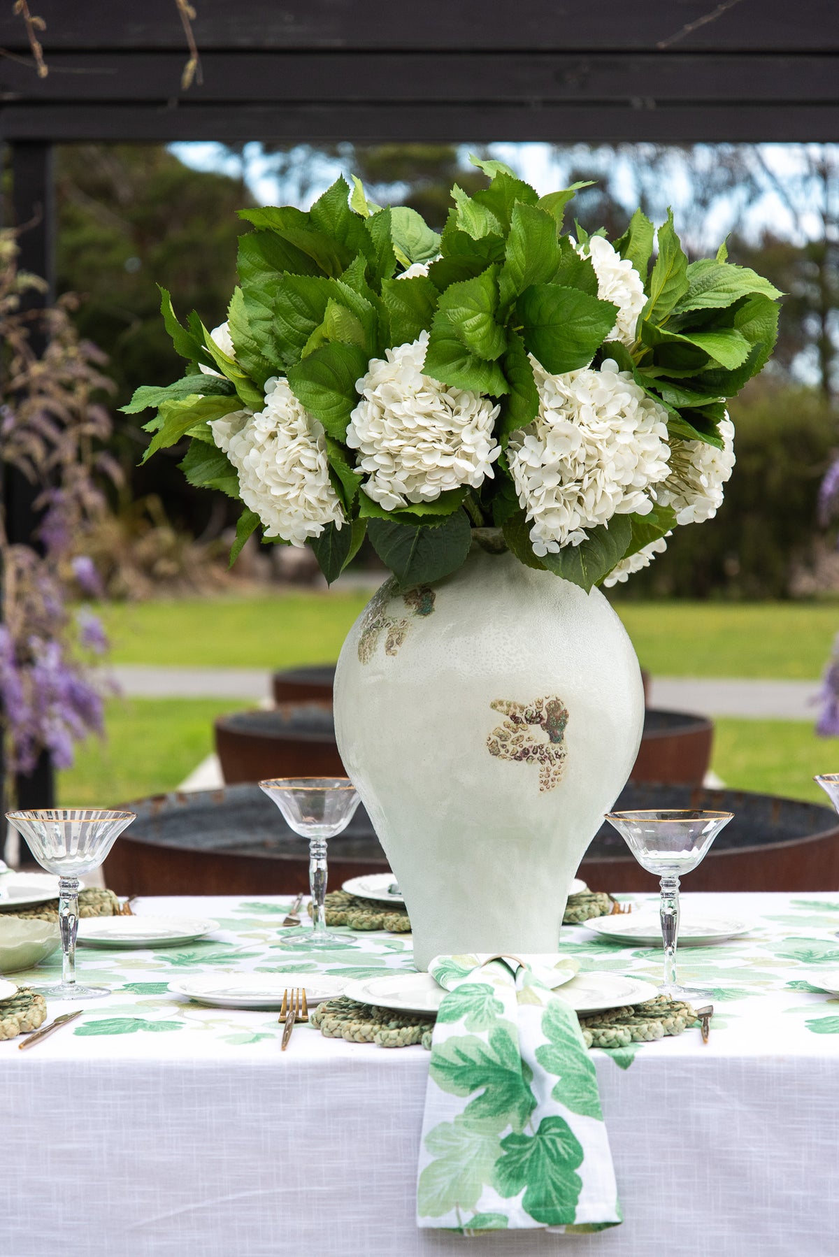 A large white ceramic vase with a green botanical pattern, filled with white hydrangea flowers and green leaves, set on a decorated outdoor table.