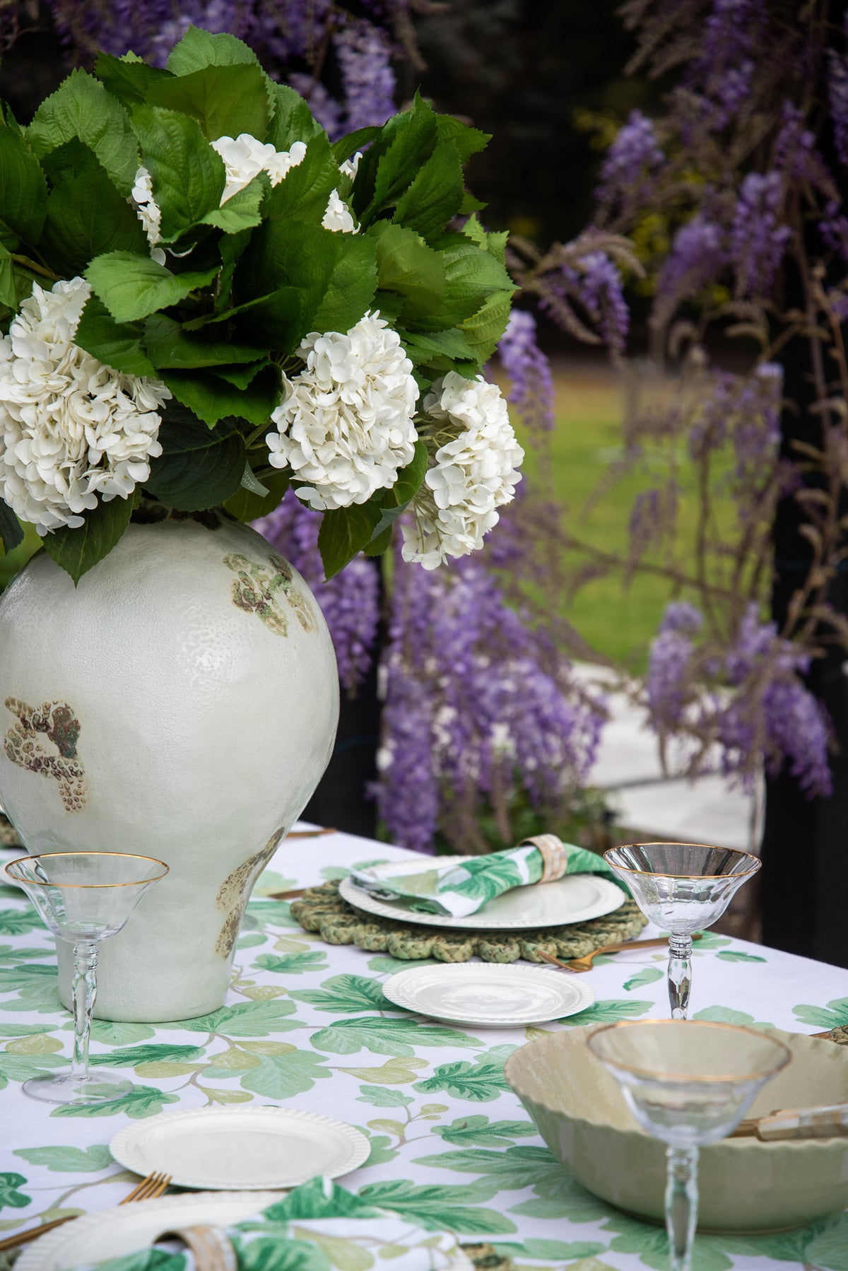 A styled outdoor table setting with a large white ceramic vase holding white hydrangea flowers and green leaves, set on a green and white patterned tablecloth with plates, napkins, and martini glasses. Purple wisteria flowers hang in the background.
