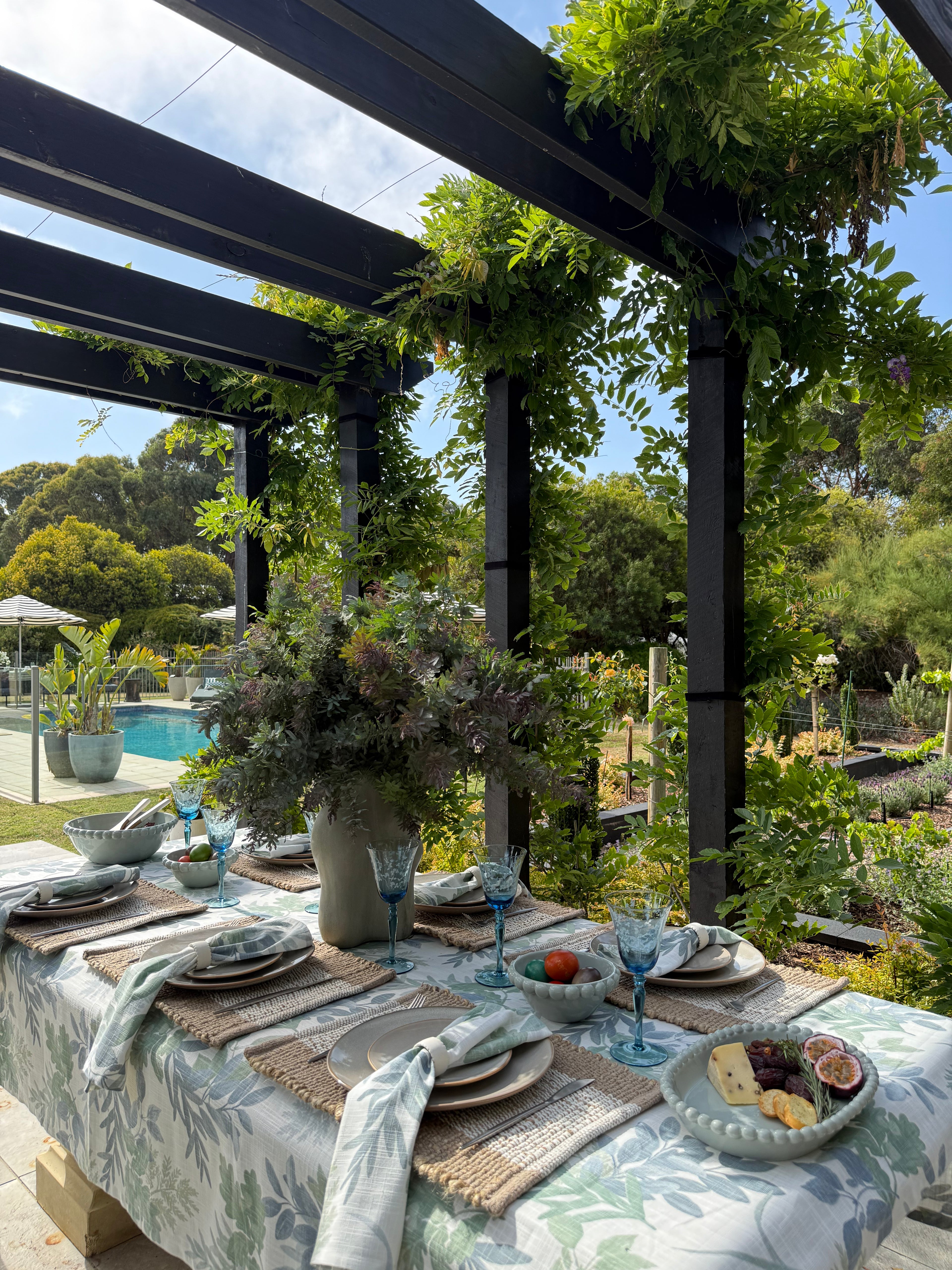 Outdoor dining setup with a tablecloth, plates, and glasses under a pergola with greenery.