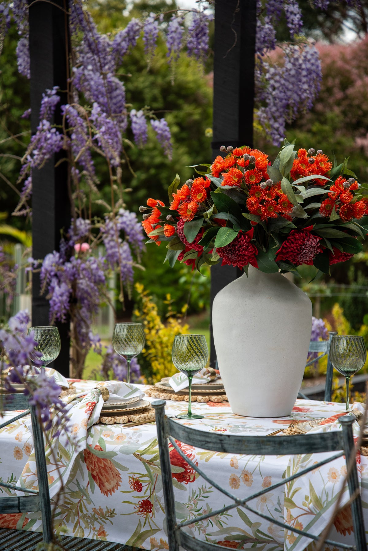 A white ceramic vase filled with a vibrant bouquet of orange and red flowers, set on a garden table with a floral tablecloth, green chairs, and a lush background of hanging purple wisteria.