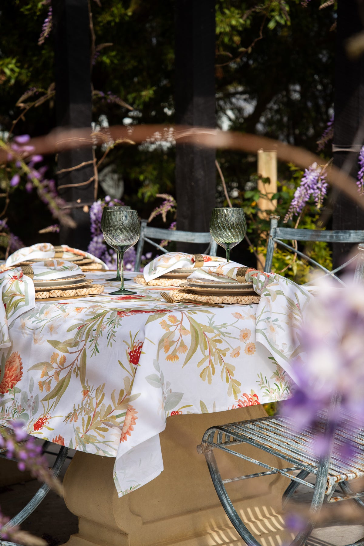 A round outdoor table set with a white tablecloth featuring a botanical floral print in orange, green, and beige. The table is styled with folded napkins, green textured wine glasses, and woven placemats, set against a background of patio chairs and greenery with purple flowers in the foreground.