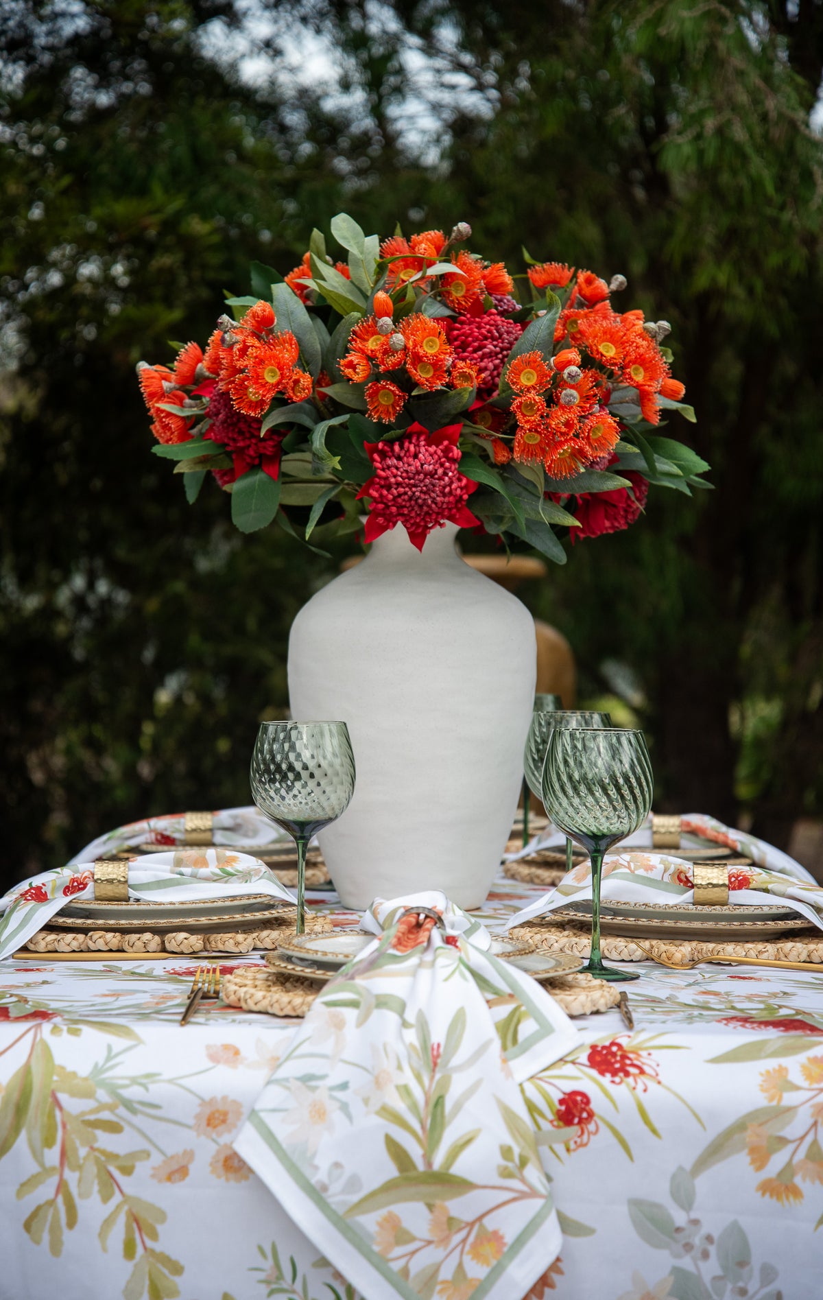 A styled outdoor table setting with a large white vase holding a vibrant orange and red floral arrangement, set against a blurred green forest background.
