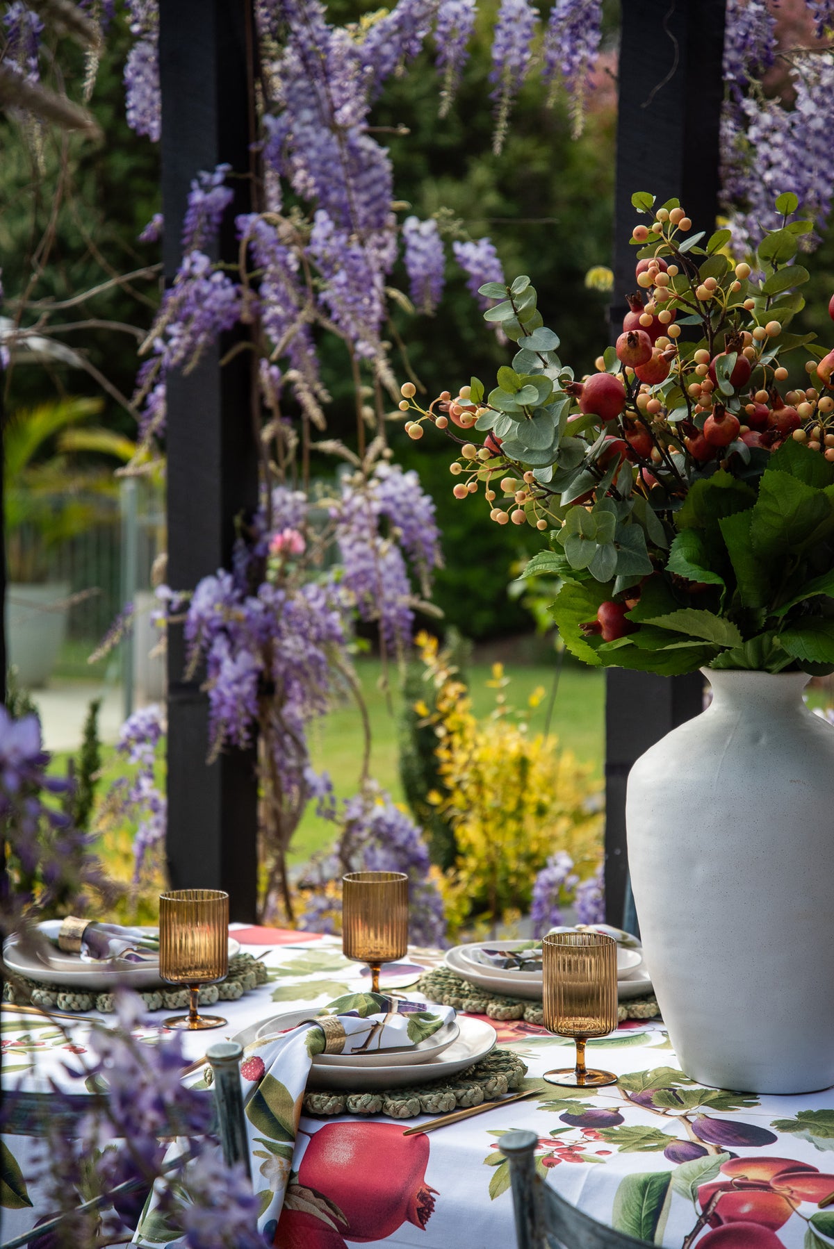 A styled outdoor dining table draped in a colorful fruit-patterned tablecloth, decorated with a large white vase of red berries and greenery, gold-toned candle holders, and surrounded by purple wisteria flowers and a garden background.