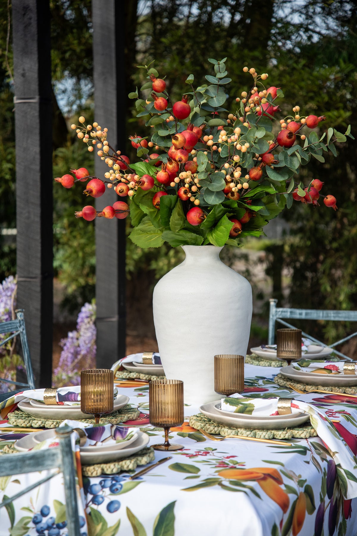 A large white textured ceramic vase filled with a colorful autumn floral arrangement of red berries, green leaves, and orange accents, set on a decorated outdoor table with a floral tablecloth and place settings.