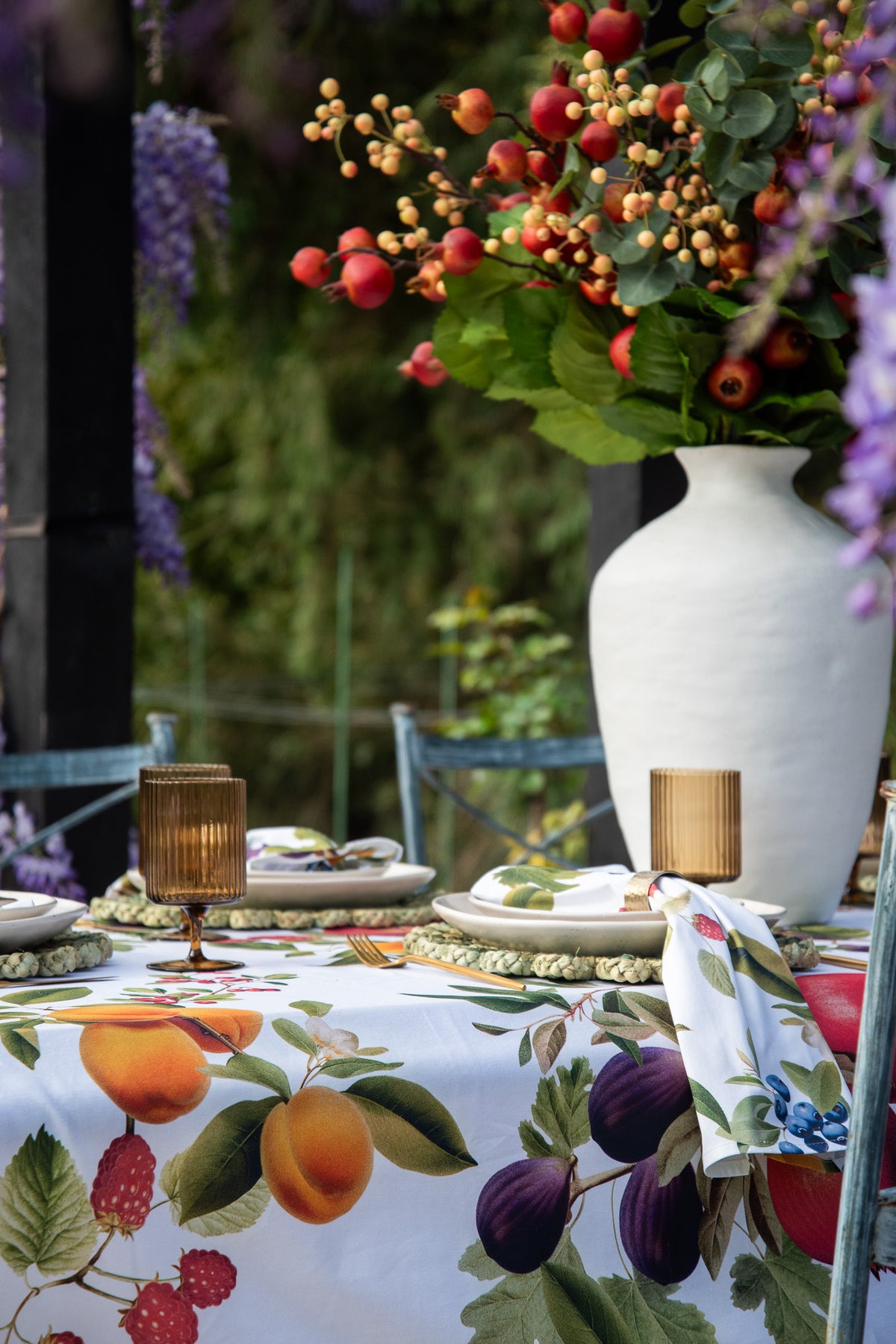 A styled outdoor dining table featuring a white tablecloth printed with a colorful fruit and berry motif, set with plates, gold-toned glassware, and a large white vase filled with a floral arrangement in a garden setting.